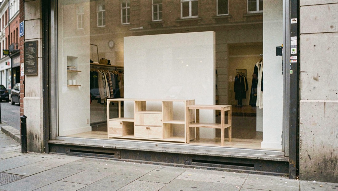 Vitrine de boutique vue depuis le trottoir, mobilier bas en bois clair et socle de présentation vide derrière la glace